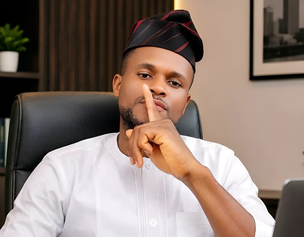 Bright Njoku at his desk, agbada and cap, finger to his lips